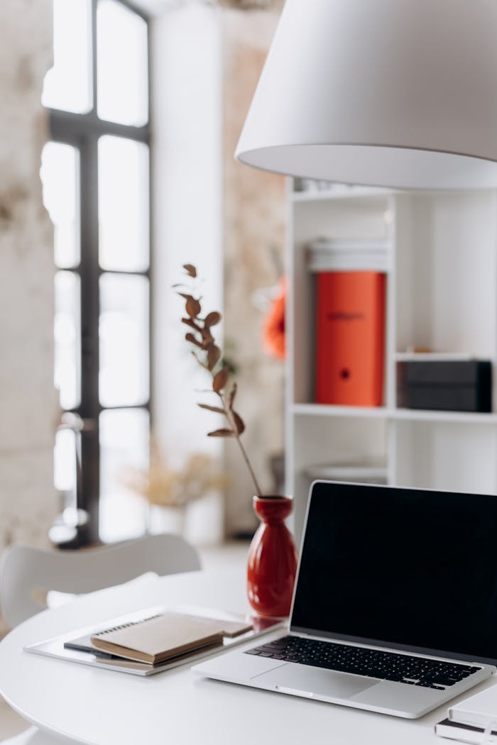 Stylish minimalist home office setup with a laptop, red vase, and decor on a white table.